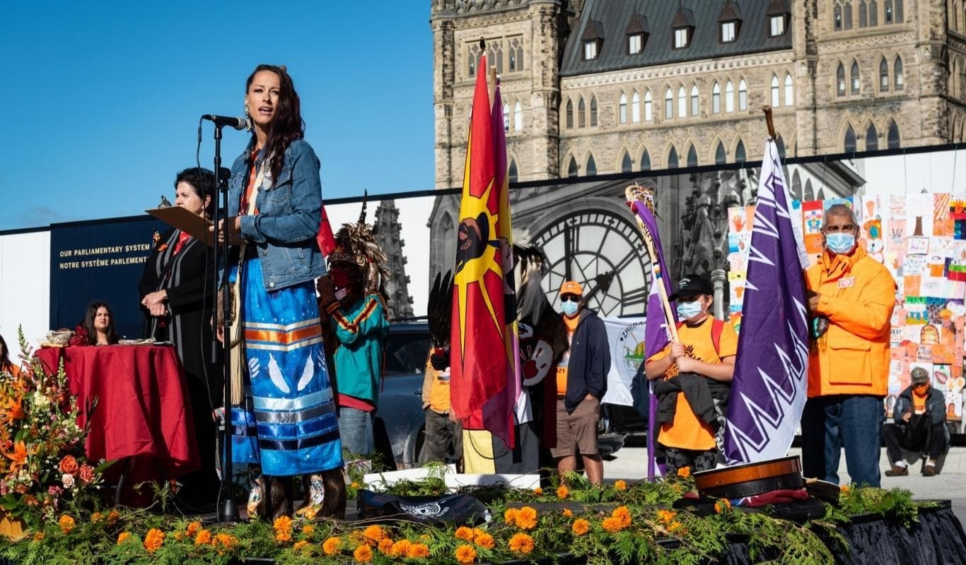 A medium-light skintoned woman with long dark hair and wearing a jean jacket, a shirt and a ribbon skirt stands on an outdoor stage in front of a microphone with Parliament Hill visible behind her. On the stage in front of her is greenery and orange flowers and behind her several medium-light skintoned people in medical masks and wearing orange hold flags from several Indigenous nations.