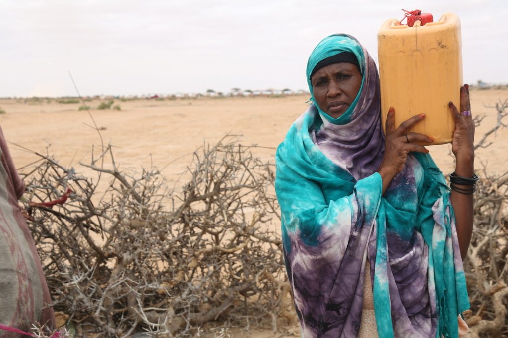 A woman in a blue and grey hijab holds a yellow water jug on her shoulder as she stands in the dry countryside.