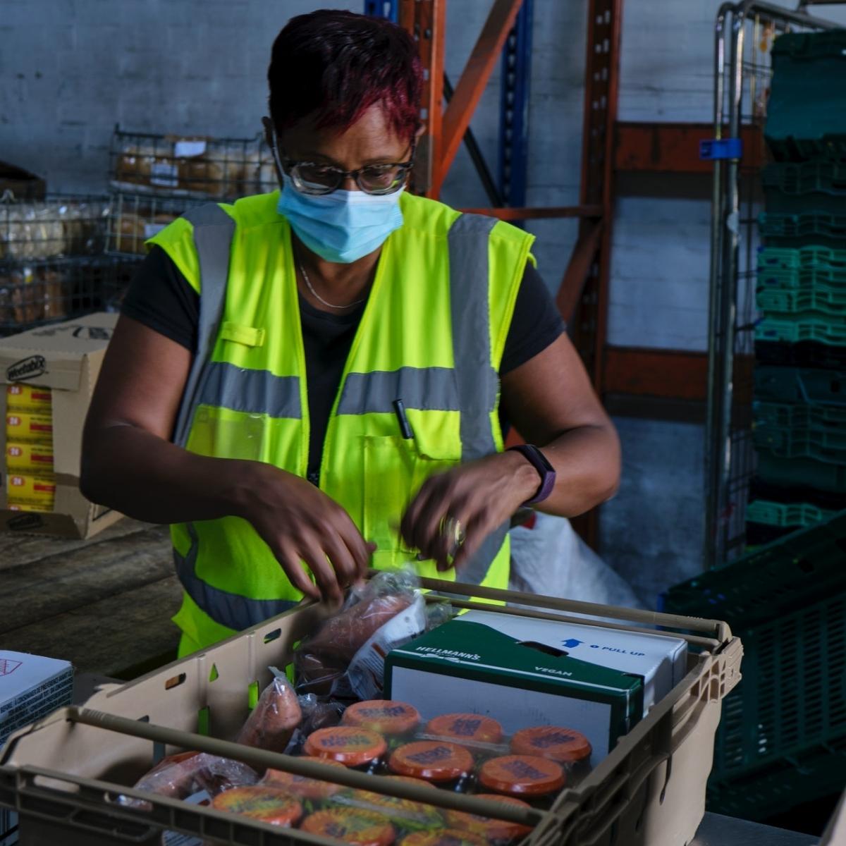 A dark-skinned woman with short, red hair wears a blue medical mask and a yellow and grey industrial vest as she packs boxes of food in an industrial setting.
