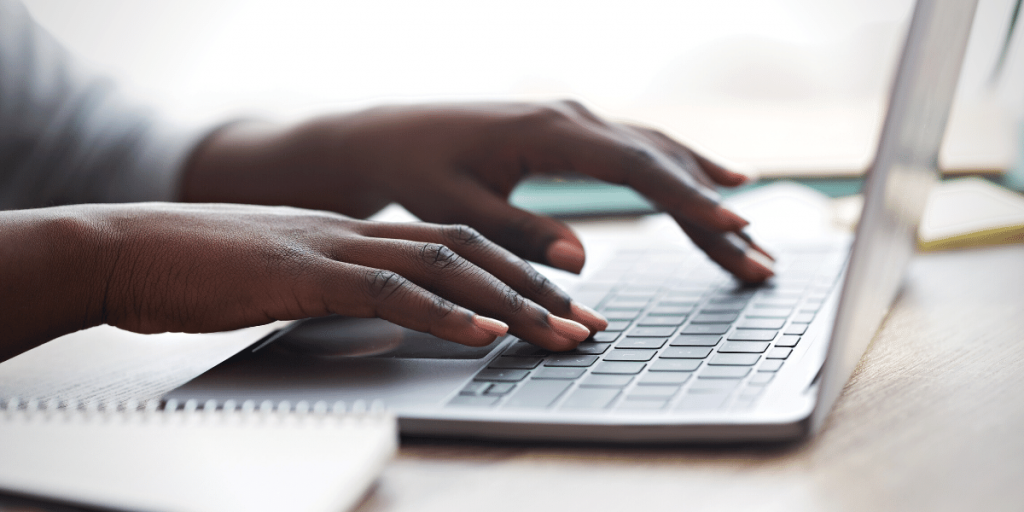 Close up of a pair of dark skinned hands typing on a laptop's keyboard.