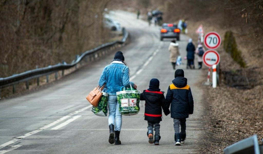 People are walking on a paved road, and two cars are parked in the distance. None of the people are facing the camera. Most of them seem to be women and children, wearing winter clothing and carrying bags. The main focus of the photograph is three people featured in the front centre, not facing the camera. A woman in blue jeans and a blue jean jacket carries a brown purse and two big reusable supermarket bags. Two boys wearing navy blue winter coats and winter boots are to her right. The shortest one holds both the woman’s hands and the taller boy’s hands.