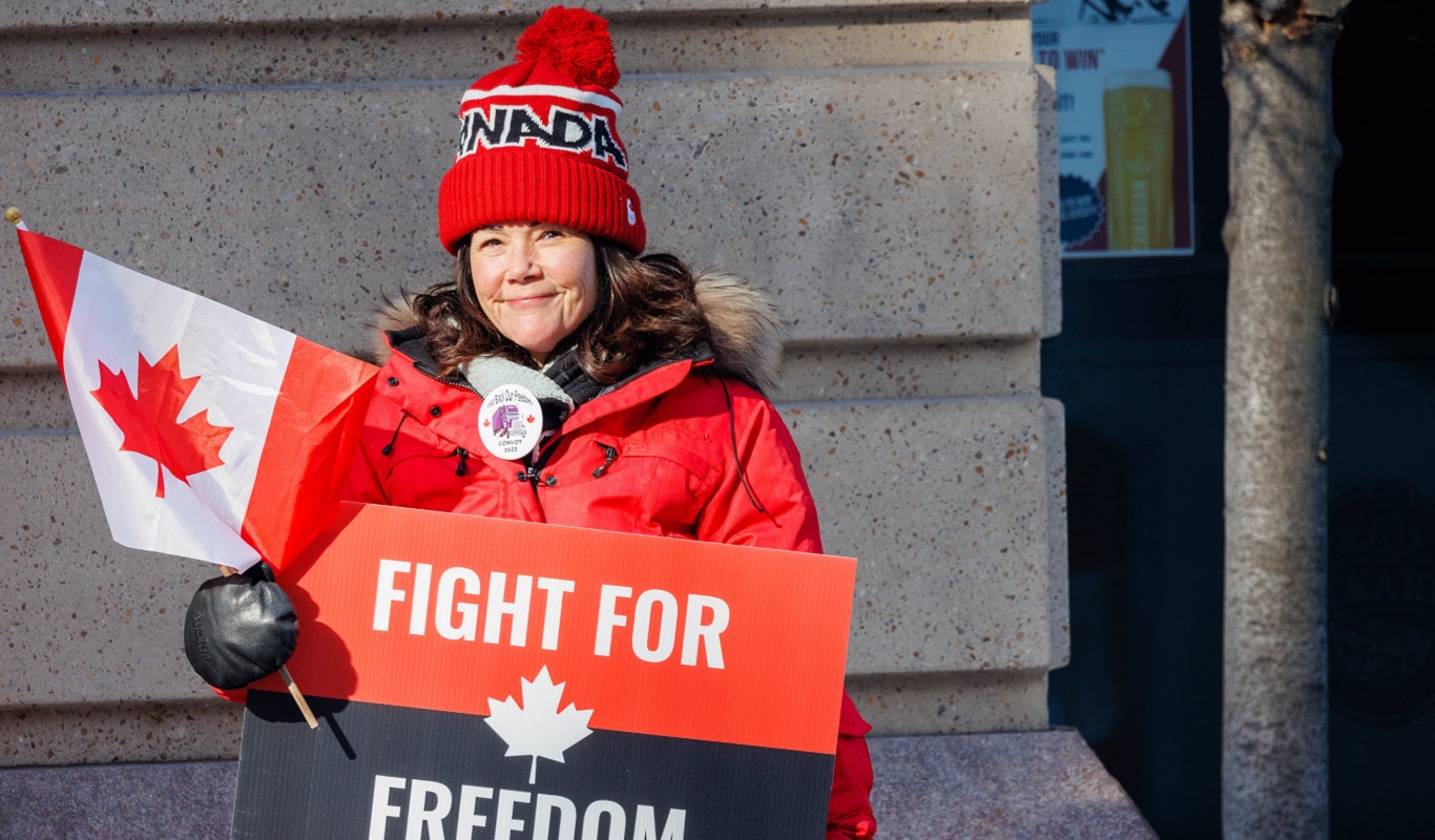 A light-skinned woman stands in front of a stone wall. She is holding a Canadian flag in her right hand and a sign that reads Fight for Freedom in her left hand. She is wearing a red jacket and a red, white and black tuque with the word Canada on it. She appears to be smiling for the camera.