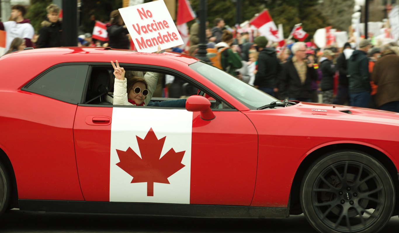 2022 Convoy Woman - shutterstock_2120035874 - 1366x800px A middle-aged light-skinned woman with dark, round sunglasses and a white sweater on gives the peace sign out of the window of the car she is riding in, which is an old red camaro painted to look like the Canadian flag. There is a crowd of protesters behind the car, and one sign is more visible than the others. It reads No Vaccine Mandate.