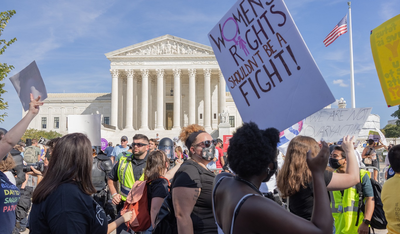 Roe v Wade protests in US - shutterstock_2051768072 -1366x800px Dozens of activists of varying genders and skin tones are gathered together. Most of them are wearing face masks and some are carrying signs. One of the most visible signs is being carried by a dark-skinned person in a spaghetti-strapped tank top, and the sign reads women's rights shouldn't be a fight.