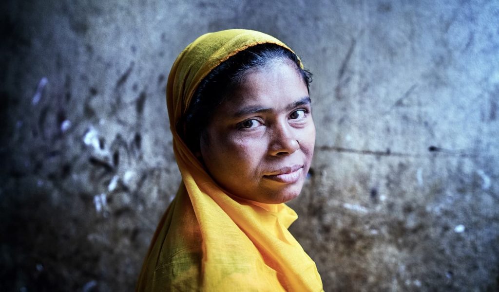 A brown skinned middle age woman wearing a canary yellow headscarf stands in front of a rough-textured concrete wall with a serious look on her face.