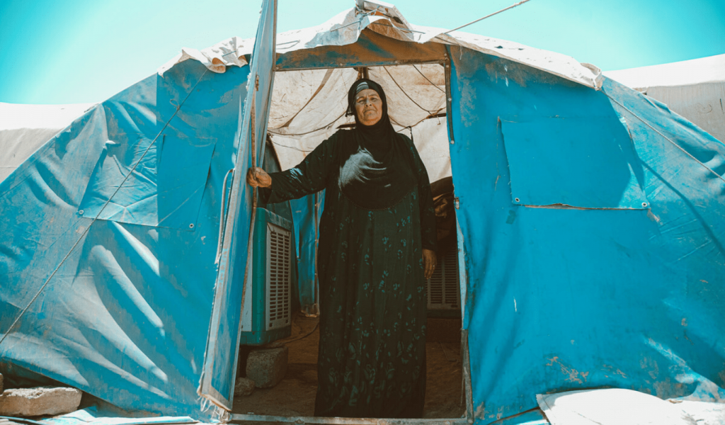 An elderly light-skinned woman stands in the doorway of her family's light blue tent. The woman is wearing a long-sleeved ankle-length dark green patterned dress and a black headscarf. Behind her, the sky is clear and bright.