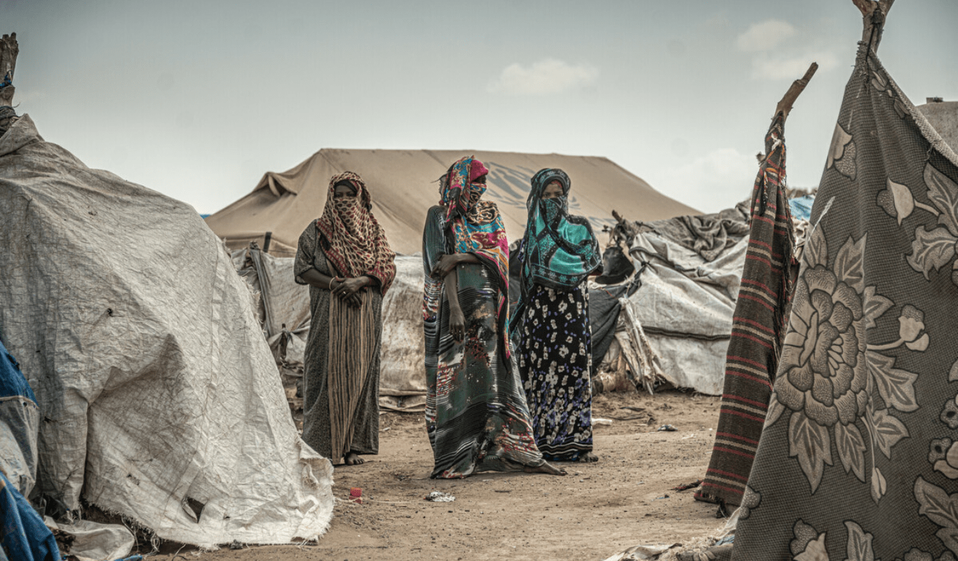 RS247015_44203_f82397c5c0872a1-scr_min Three women wearing colourful hijabs stand outside, surrounded by white, makeshift tents against a cloudy sky. Beneath them the ground is brown.