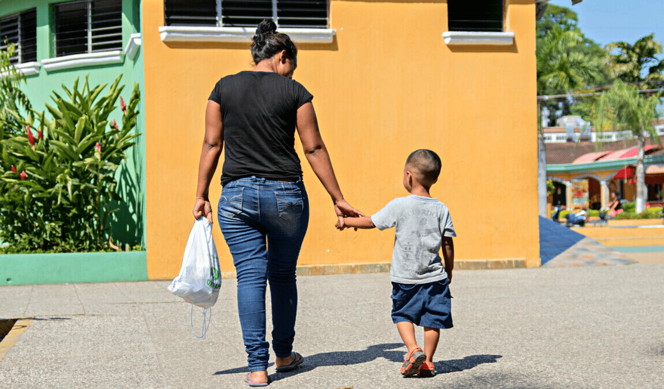 RS250304_54111_0146a5b77626d73-scr-min A woman wearing jeans and a black shirt carries a white plastic bag with food items while holding the hand of a small boy. Both are outside, passing through yellow and green buildings and to our left, is a park.