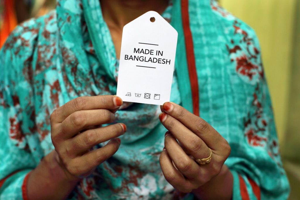 Close up of a woman wearing a turquoise long sleeve shirt with red flowers holding a tag that reads "Made in Bangladesh." The photo doesn't show her face, just her upper body.