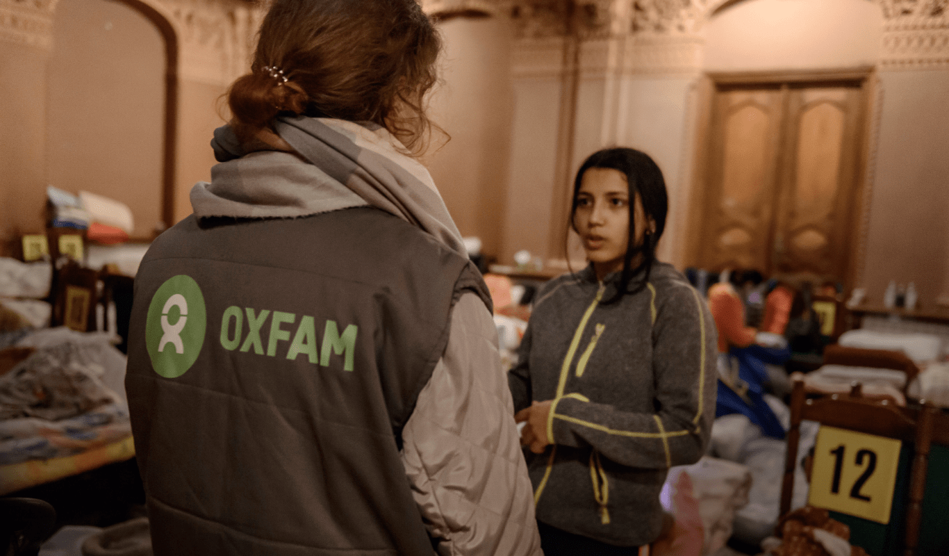 Two women talk, one faces directly the other woman who is not facing the camera. They're both wearing sweaters. One of them wears an Oxfam branded vest.