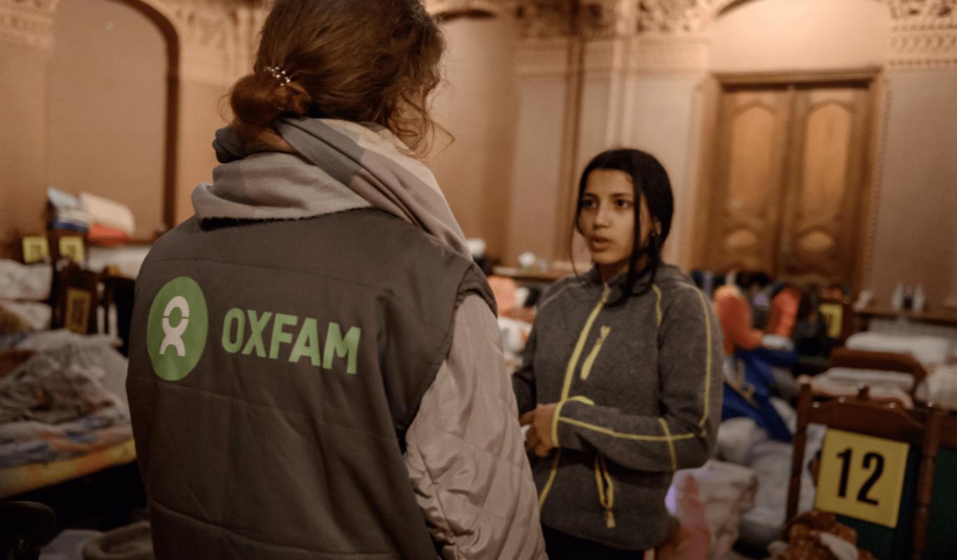RS340673__OBE6291-scr_min Two women talk, one faces directly the other woman who is not facing the camera. They're both wearing sweaters. One of them wears an Oxfam branded vest.