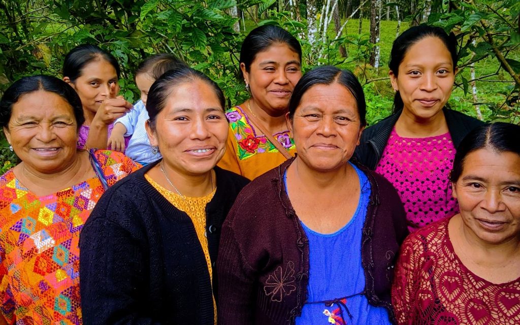 A group of seven Indigenous women wearing a mix of traditional clothes and non-traditional blouses are smiling and looking directly at the camera. They're standing outside with luscious green trees behind them.