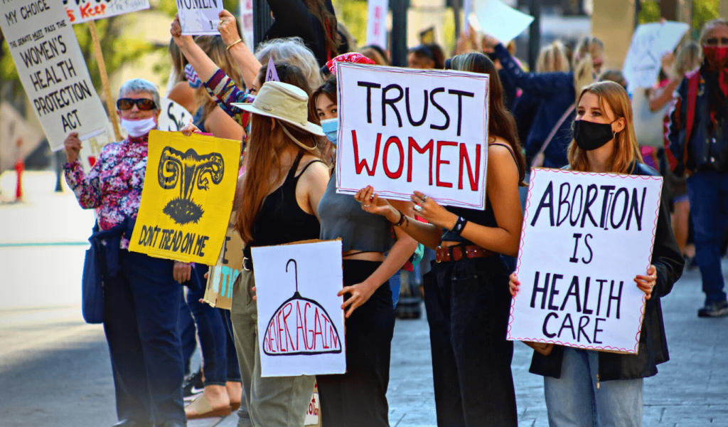 A group of women ranging from different ages holding protest signs that say messages like, abortion is healthcare, trust women, a depiction of an uterus with the phrase underneath that says, don't tread on me.