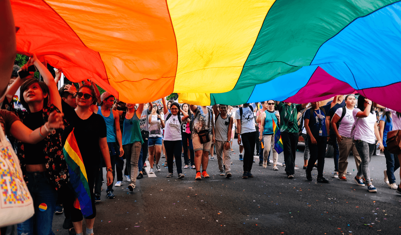 A group of people from different genders and ages walk on the street under a giant pride flat, which some of them are carrying at the sides.