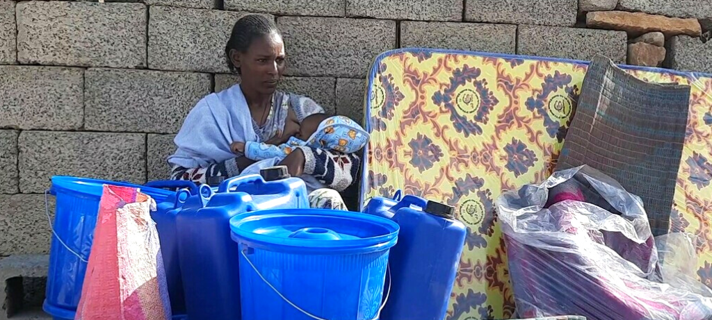 A woman is sitting down against an adobe wall, beside some flower-patterned mattresses. She is breasfeeding her baby who is wrapped in blue blankets. There are blue plastic bins in front of them.