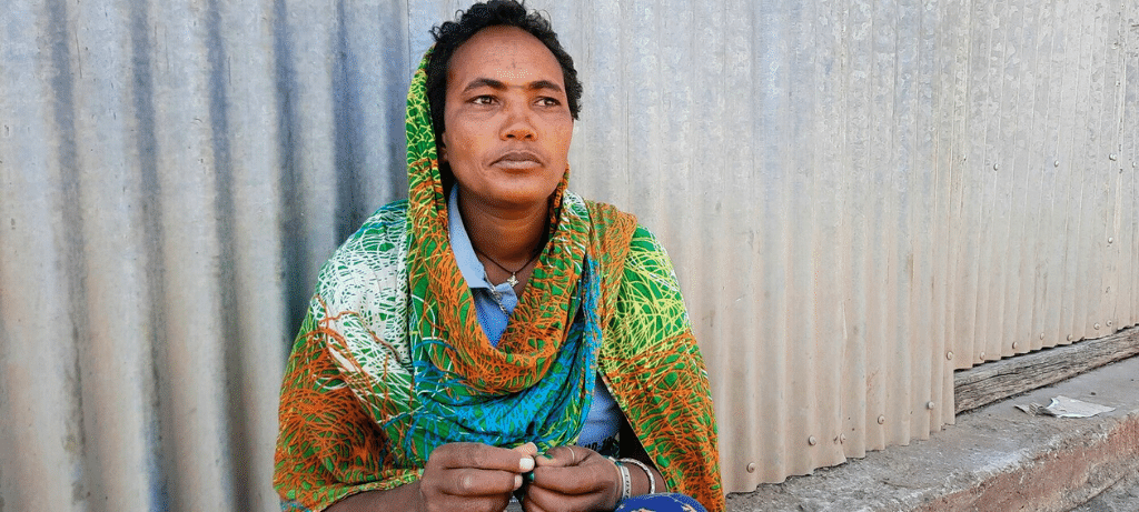 A woman wearing a colourful orange, white and green headscarf is sitting on the floor, against a corrugated metal sheet, looking at the distance, not smiling.