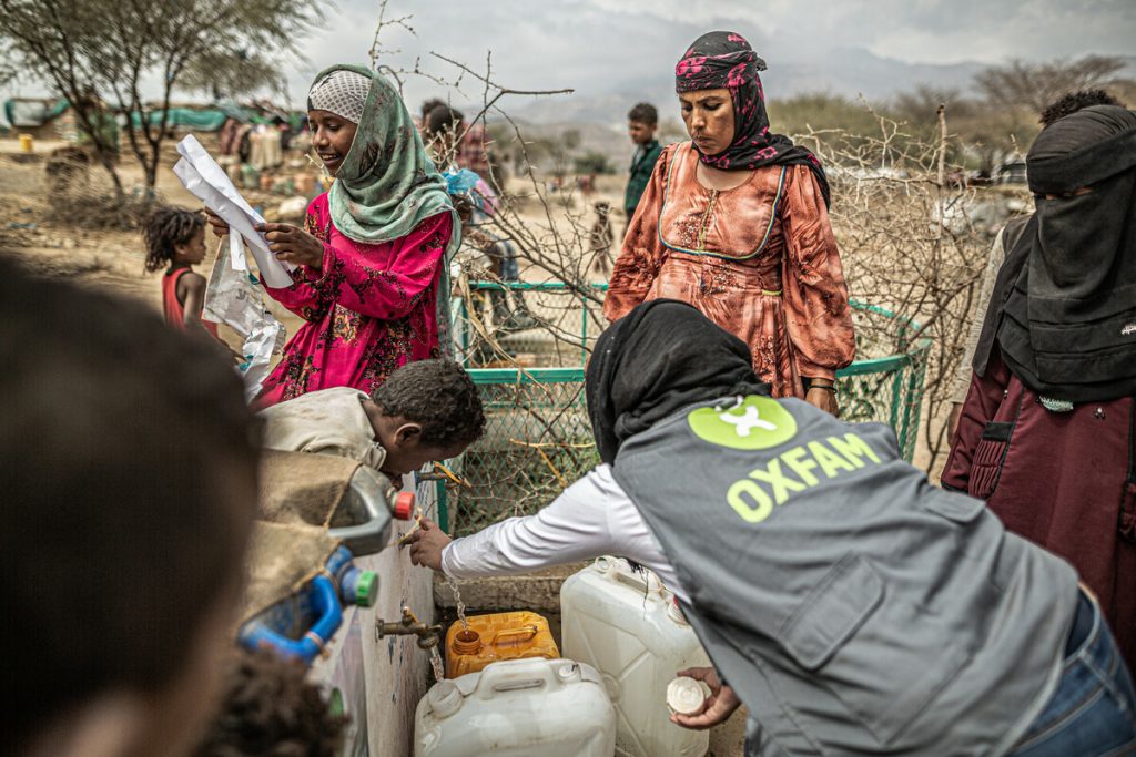 A group of dark-skinned women wearing head scarves and long dresses gather around an outdoor water distribution point. A female humanitarian worker wearing a grey vest with a green Oxfam logo fills jerry cans with water.