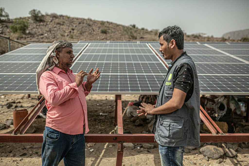 A young man wearing a grey Oxfam-branded vest faces another man wearing a pink shirt. Both stand outside in front of solar panels.