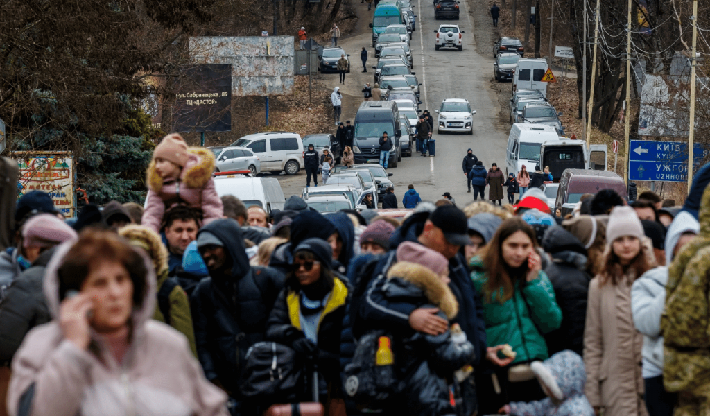 A group of people of diverse ages, genders and races wearing winter clothes stand in a crowd on a highway, several of whom are on their cell phones. Behind them are dozens of vehicles. Some parked at the side of the road and some are at a standstill on the road itself. There are several roadside signs. Some are billboards and some are directional signs.