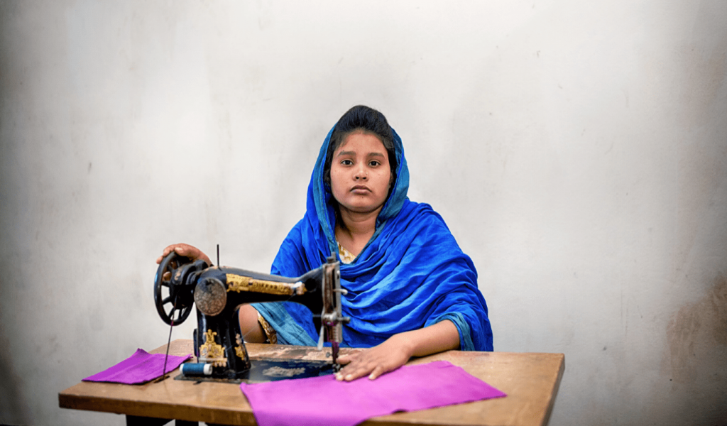 Portrait of a young Bangladeshi woman wearing a blue headscarf, looking directly at camera, not smiling. She's sitting behind a brown and black sowing machine, working on a piece of bright pink clothing in a room with white walls.
