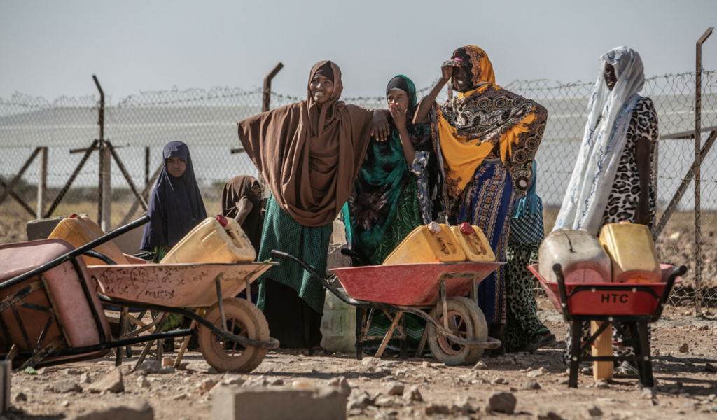 Five women and a young girl wearing colourful headscarves and long skirts stand by red wheelbarrows with yellow jerrycans waiting to collect water.
