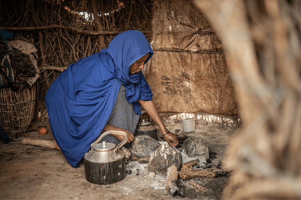A woman wearing a blue headscarf covering her head and half of her upper body kneels by a makeshift stove made of rocks inside her hut made of sticks, to boil a kettle.