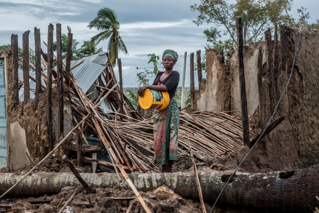 A Black woman holding a yellow bucket stands outside her destroyed house surrounded by palm trees against a cloudy sky.