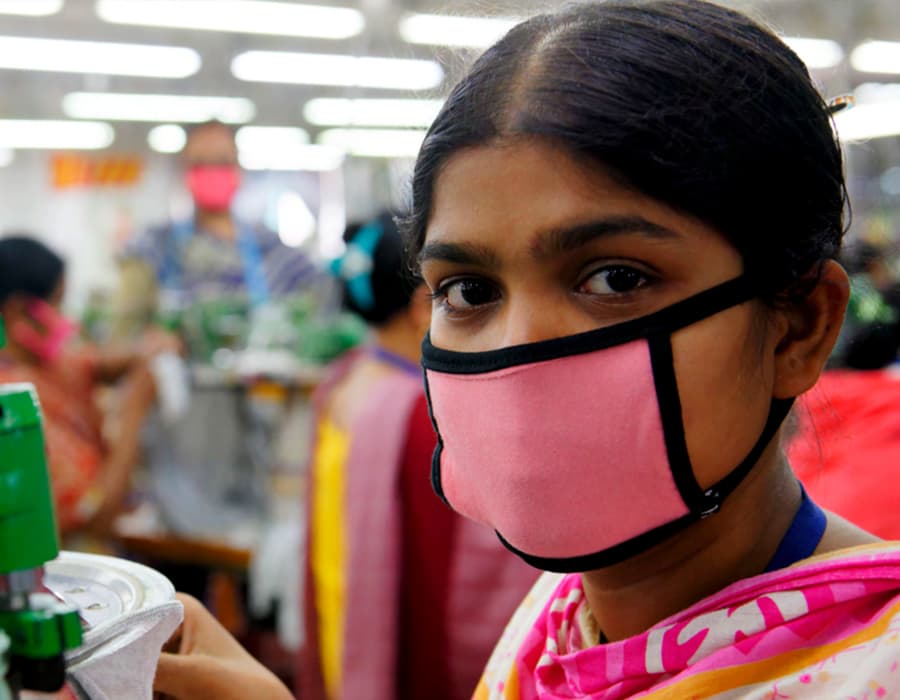 A garment worker with black hair and a pink face mark looks at the camera while working on a sewing machine in a clothing factory
