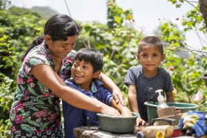 A woman and two boys wash their hands outside surrounded by trees.