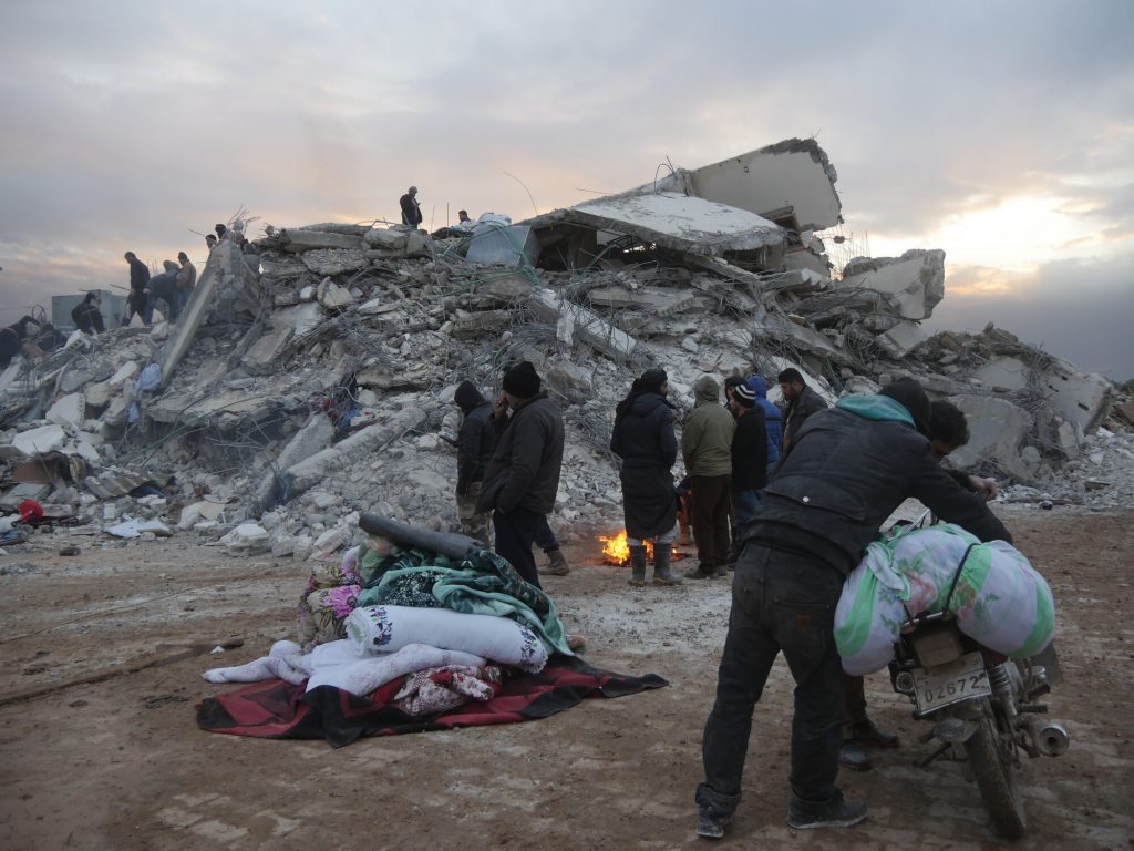 Several men are standing in front of a large pile of building rubble and around a small fire. There are large blankets rolled up on the ground in a pile and one man is holding a bike.