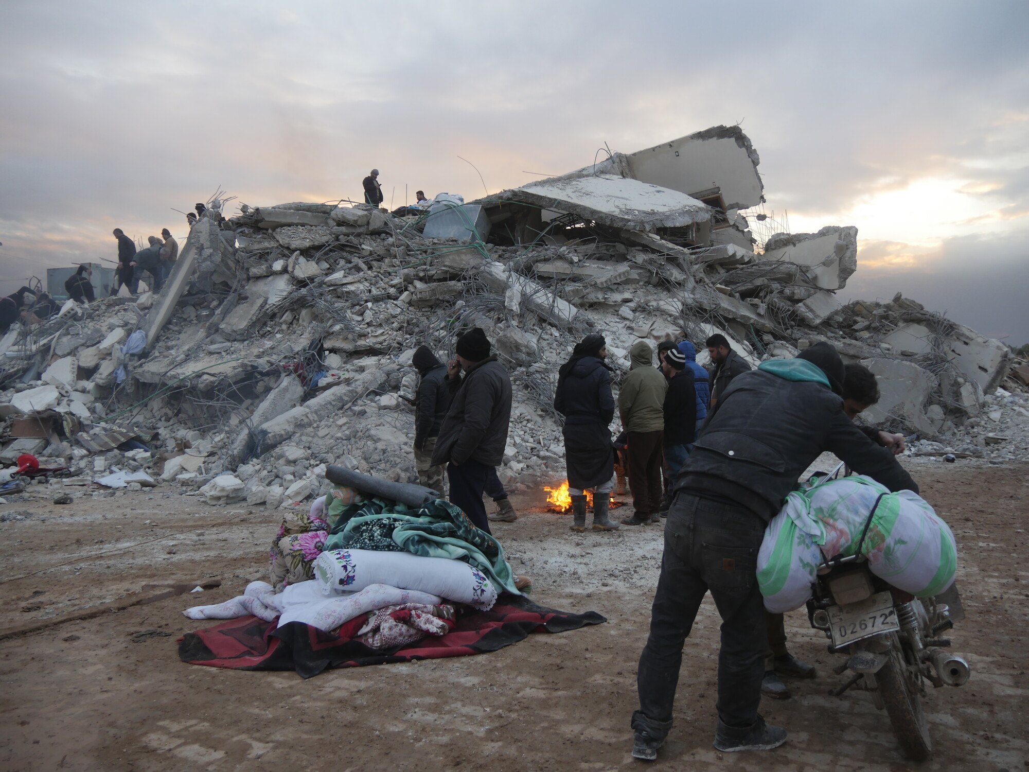 Rescue operations underway in north-west Syria, 7th February 2023 Several men are standing in front of a large pile of building rubble and around a small fire. There are large blankets rolled up on the ground in a pile and one man is holding a bike.