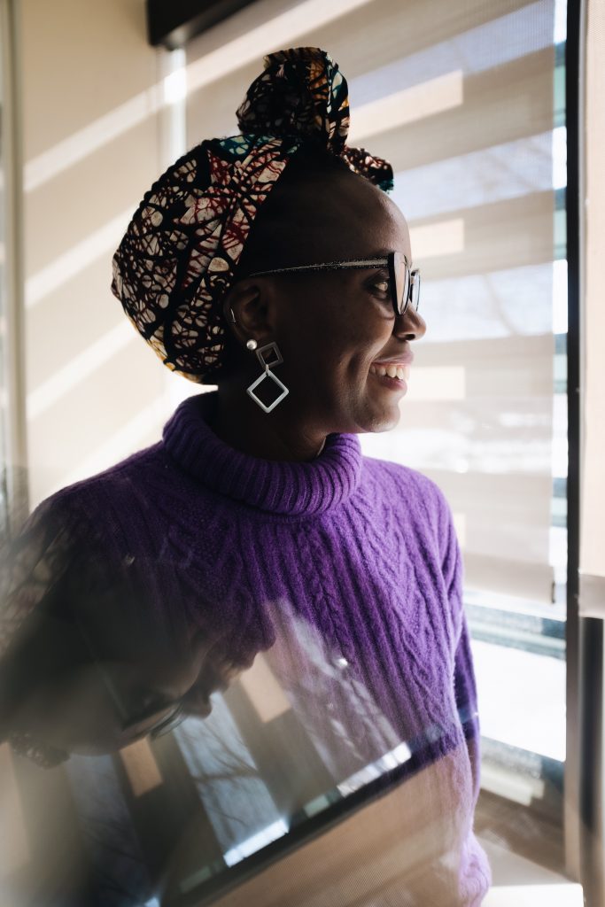 The profile of a black woman smiling. She is wearing a purple sweater, glasses and a headscarf.