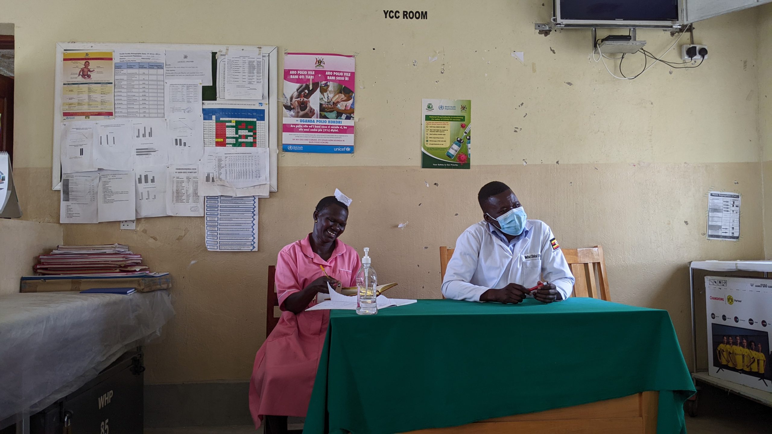 A black woman wearing a pink nursing uniform dress sits next to a black man wearing a mask and doctor's coat.