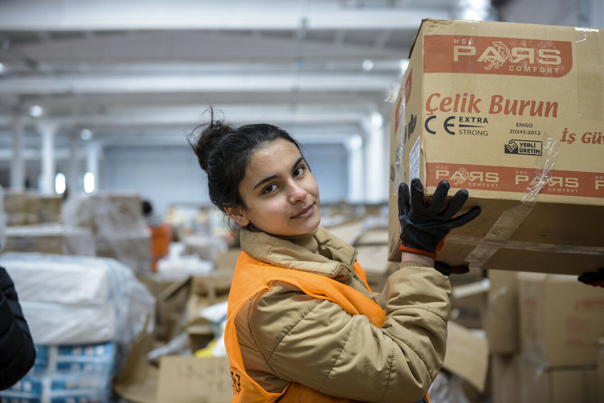 Volunteer Berfin Akdeniz A young woman with dark hair, wearing brown puffy coat and vest is looking at the camera as she lifts up a large cardboard box.