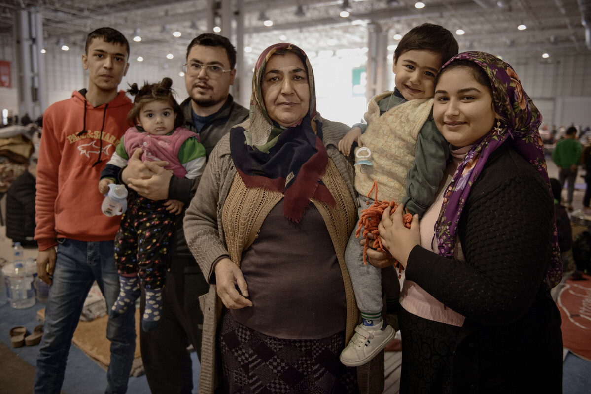 Two men stand, one holding a baby, beside an older woman and a younger woman holding a toddler in a refugee centre.