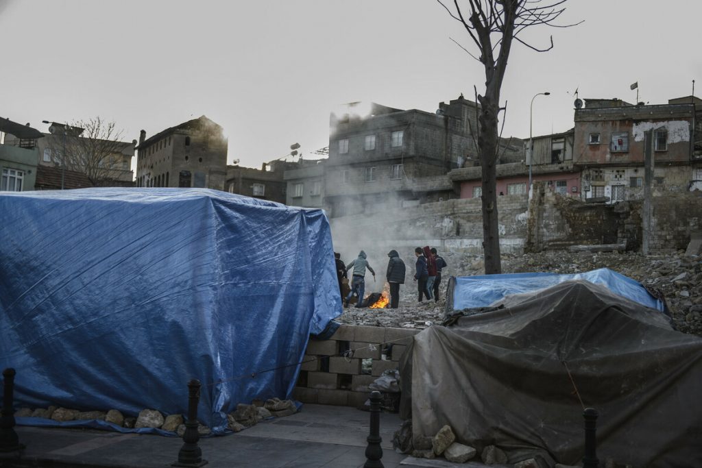 A large blue plastic tent sits on a road of rubble surrounded by fallen buildings with a group of men stand off in the distance around a fire.