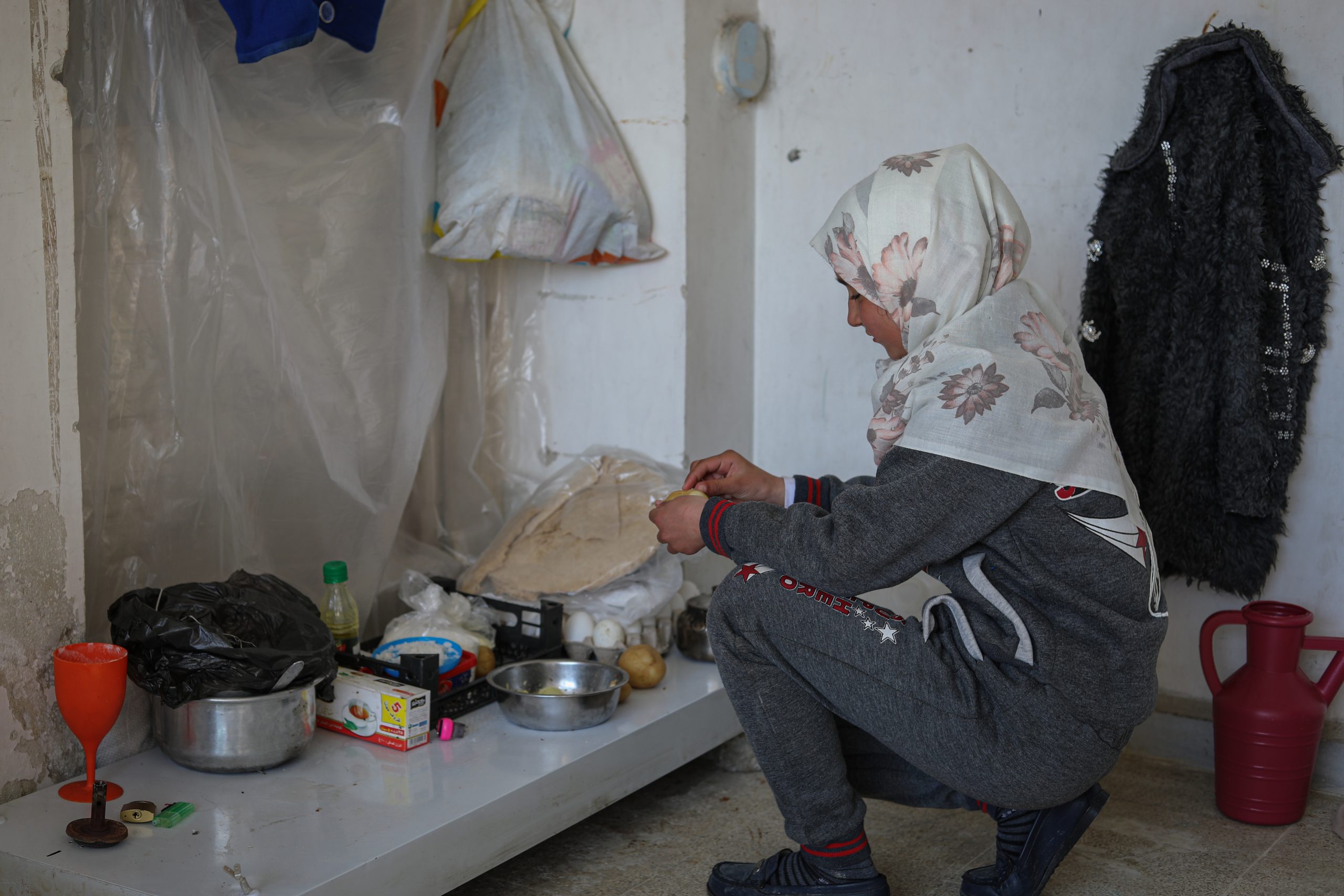 Samira_Syria_earthquake A young girl iin a grey sweatsuit and floral head covering is crouched in a corner in front of a low grey table. The table has pots, plastic coverings, a plastic bottle, a red wine glass. The girl is making something.