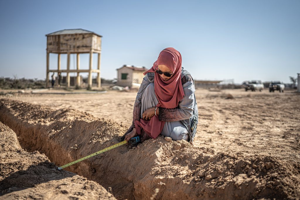 A woman wearing a red headscarf and blue clothing kneels to measure water levels of a channel. She's surrounded by dry, erosioned ground.