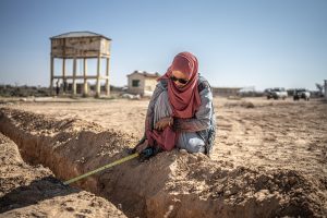 A woman wearing a red headscarf and blue clothing kneels to measure water levels of a channel. She's surrounded by dry, erosioned ground.