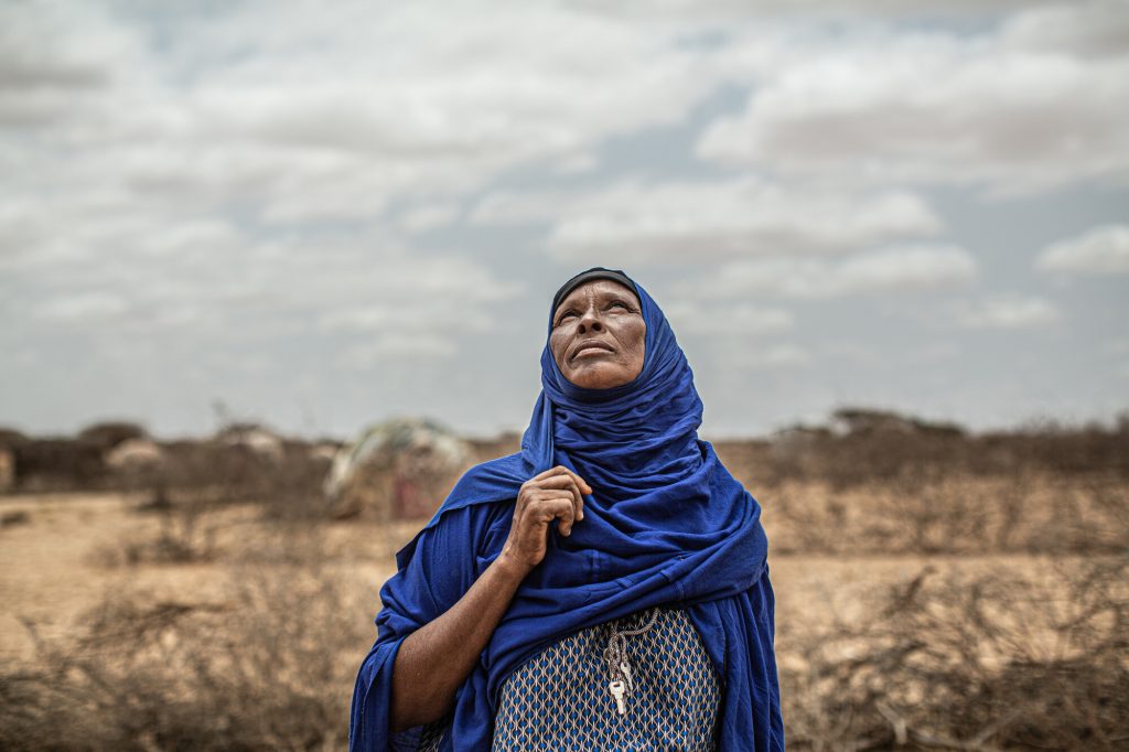 A woman is standing a barren, arid area, looking up in the sky and is wearing a blue head covering and black and white checked shirt.