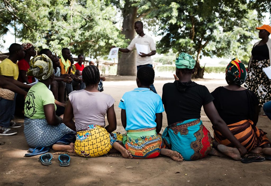 A group of people, primarily Black women wearing colourful clothing, giving their back to the camera, sit outside, surrounded by trees. They listen to an instructor who stands before them, presenting a sheet of paper displaying an image.