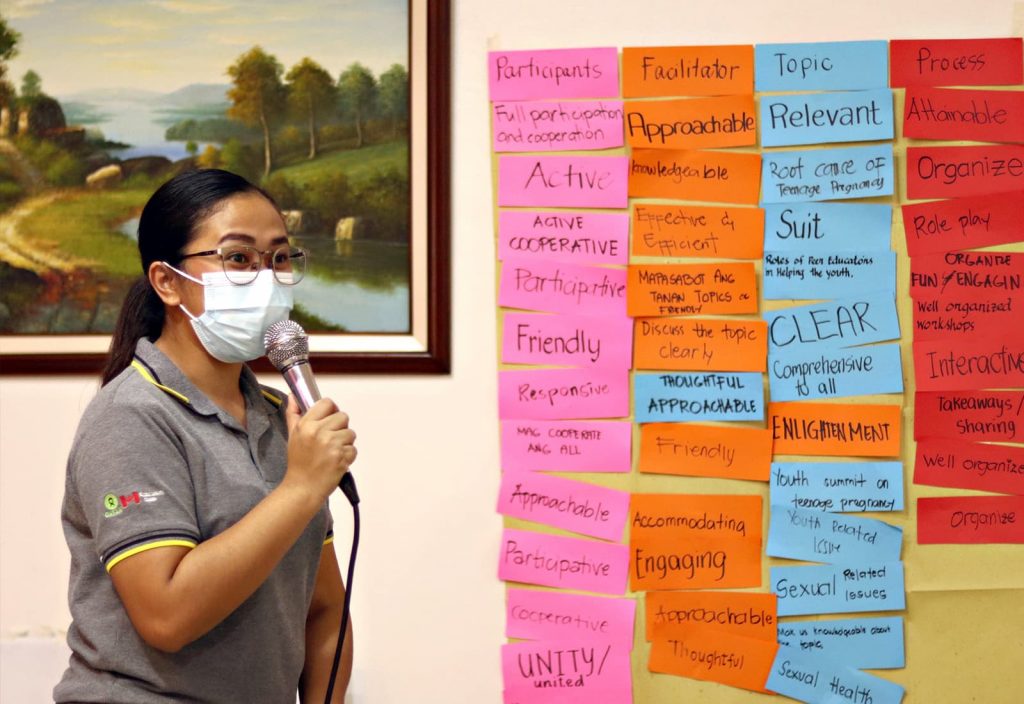 A young woman from the Philippines wearing a grey shirt and blue facemask holds a microphone while standing beside a wall covered in Post-it notes of various colours. The handwritten notes on the wall are related to reproductive health and services.