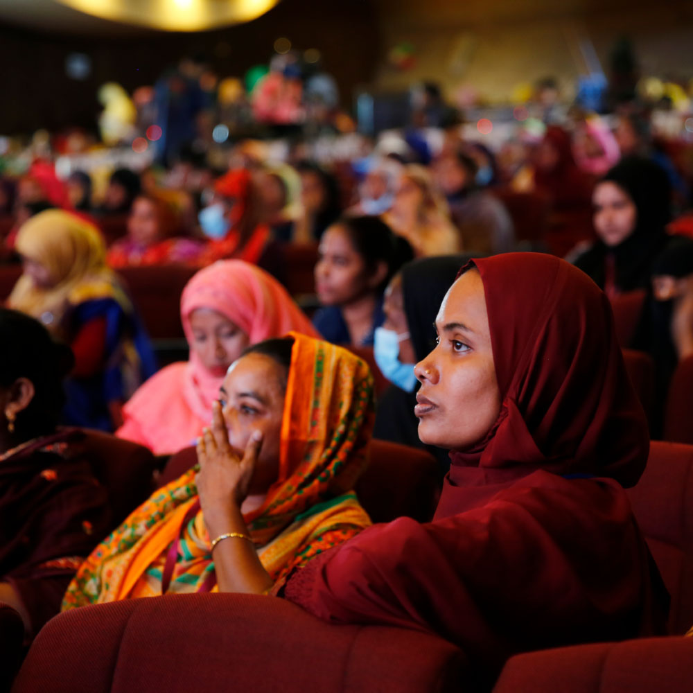 GenderJust_Hero1 A woman sits in an auditorium and listens to a speaker on stage.