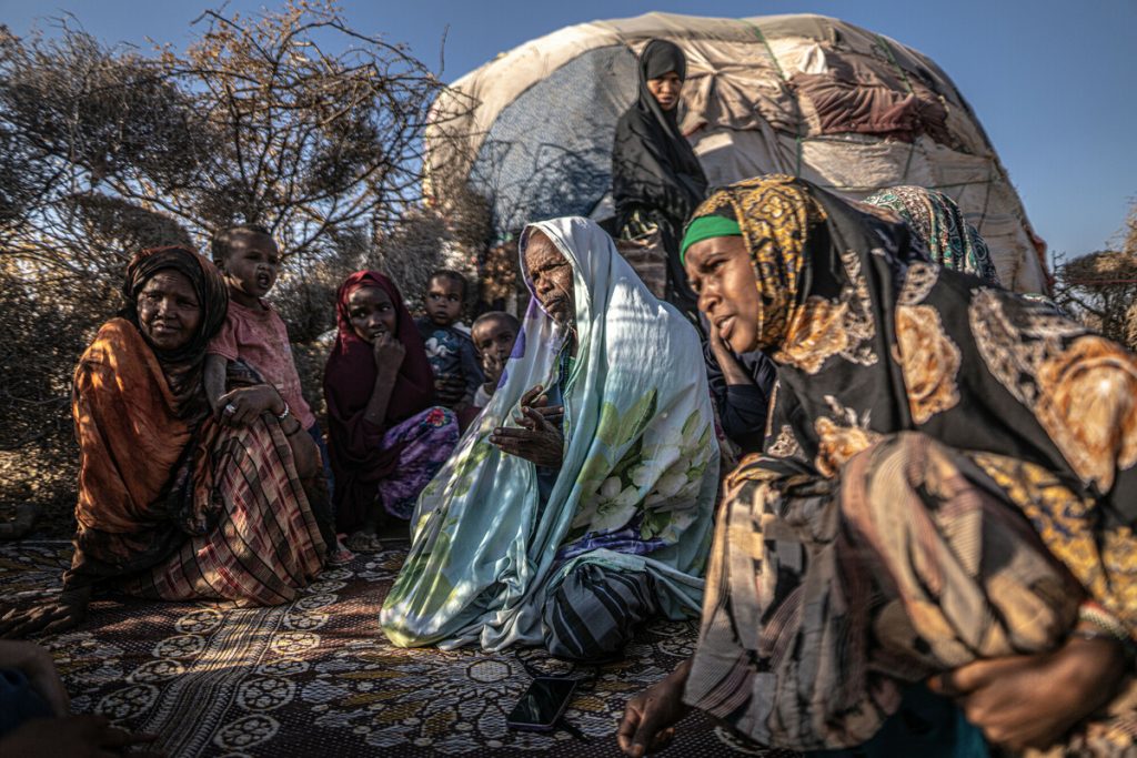 A group of women and children sit around an older man. They're sitting in the open, with a makeshift tent behind them. They wear colourful clothes and headscarves.
