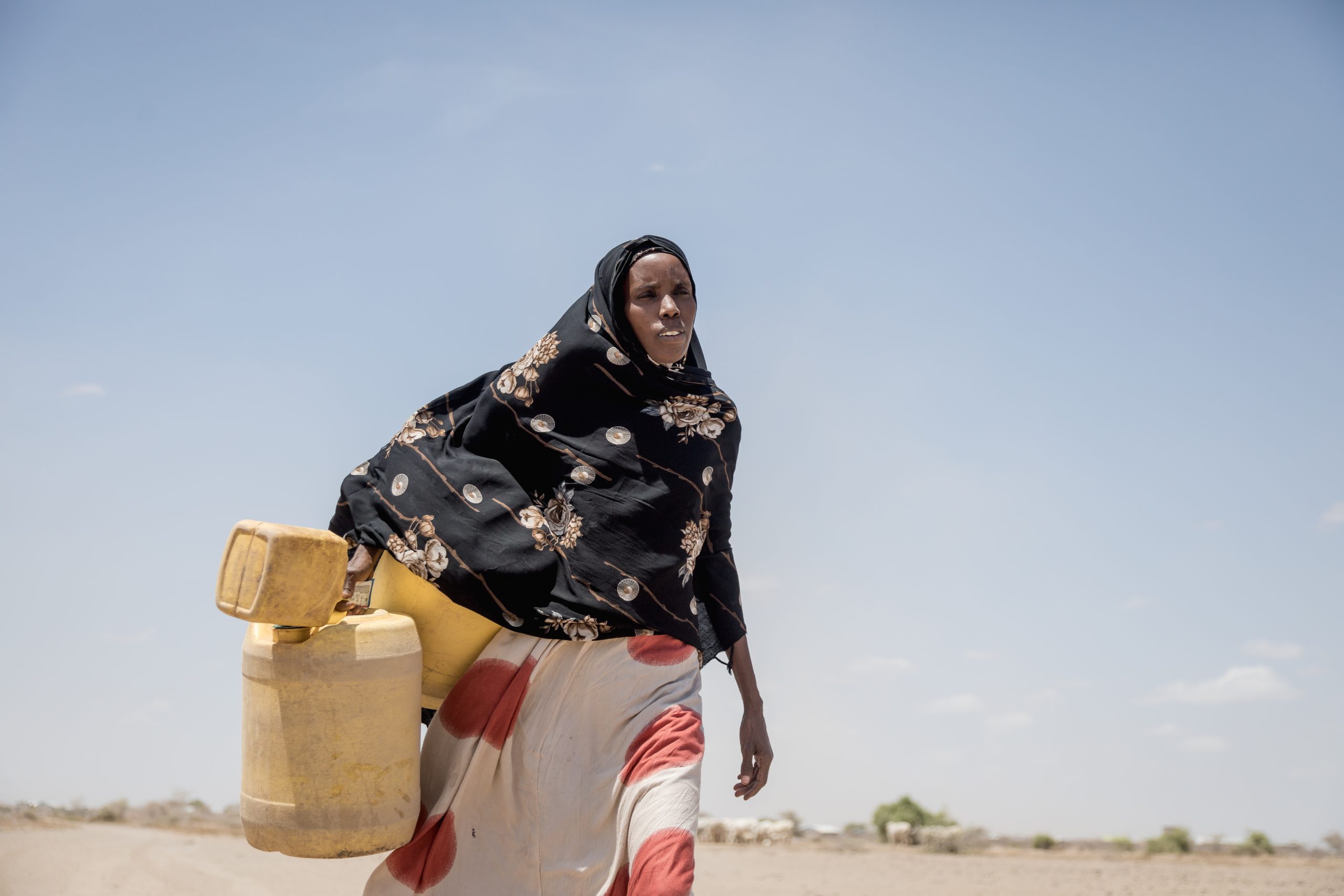 A black woman is walking in a desert-like area carring 3 empty plastic jugs of differing sizes. She has a black floral piece of material covering her head, shoulders and torso and is wearing a long white skirt with large red circles on it.