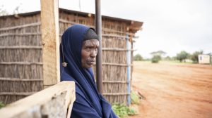 An older black woman stands sideways looking off in the distance and leaning against a wooden post in front of a thatched structure. Her head and body are fully covered by navy blue material.