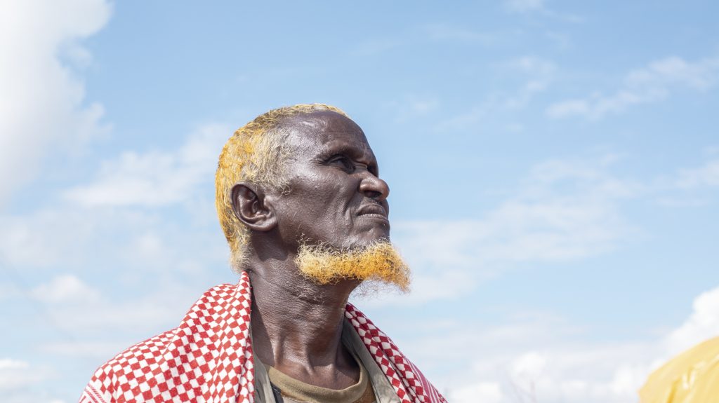 An older black man stands showing his profile from the shoulders up. He has yellow hair on his head and chin and has a red and white checked piece of fabric draped around his shoulders.