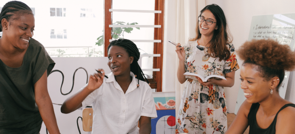 Four racially diverse women are gathered around a wooden table. Two of them are seated while the others stand, each smiling. They hold pens and notebooks and are engaged in an animated conversation.