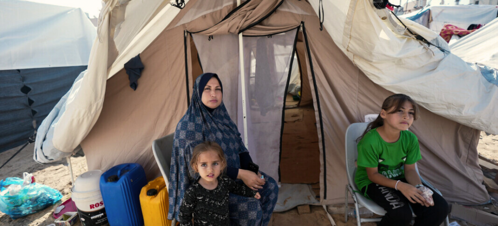 A middle-aged woman wearing a blue headscarf and two young girls with solemn faces sit outside a large white tent.