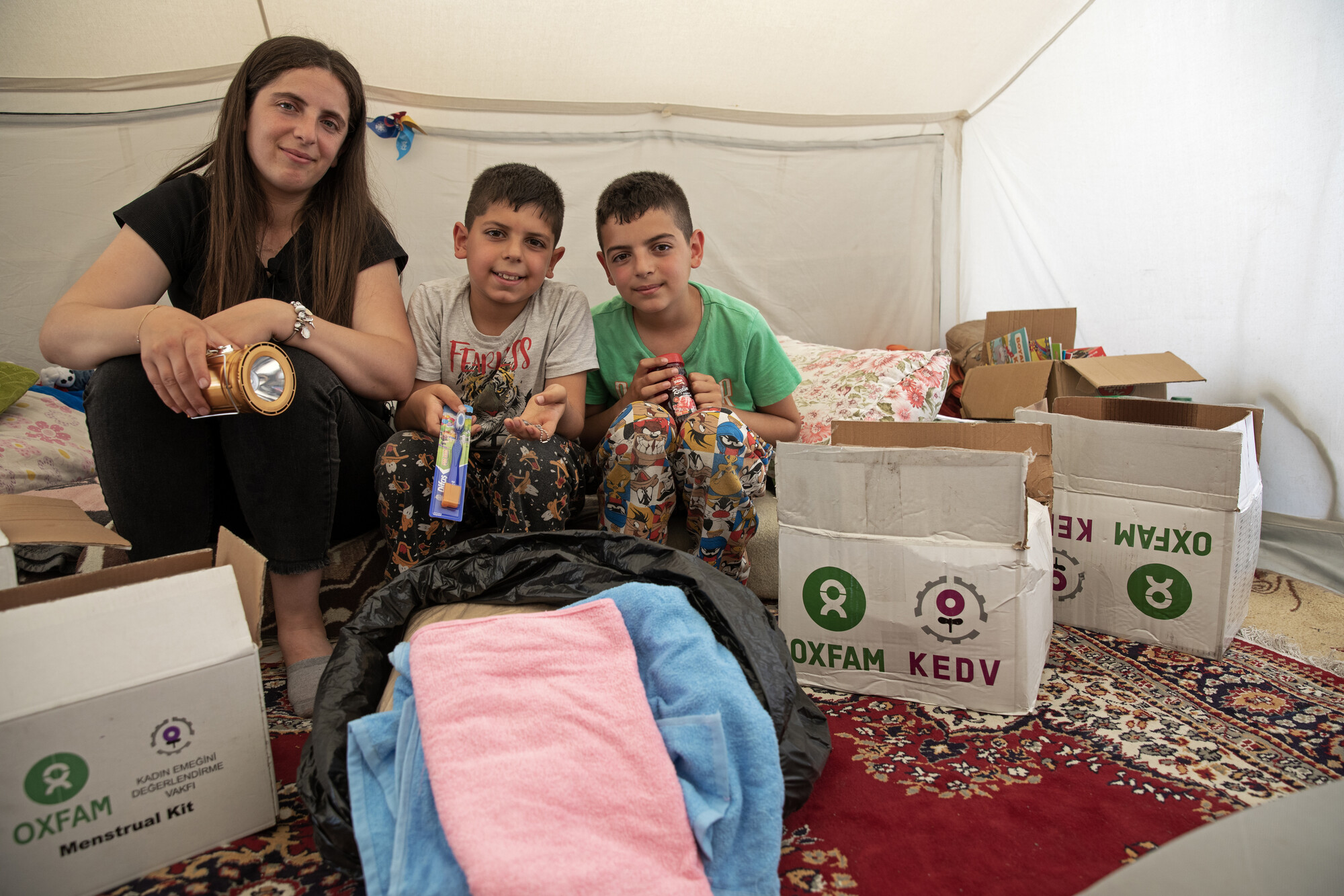 Mother and her two young sons are sitting in their tent. Surrounded by boxes of materials provided by Oxfam and KEDV. They are smiling and seem happy and grateful to have made it to a safe area. The little family is hugging and playing around while unboxing the supplies and goods boxes.